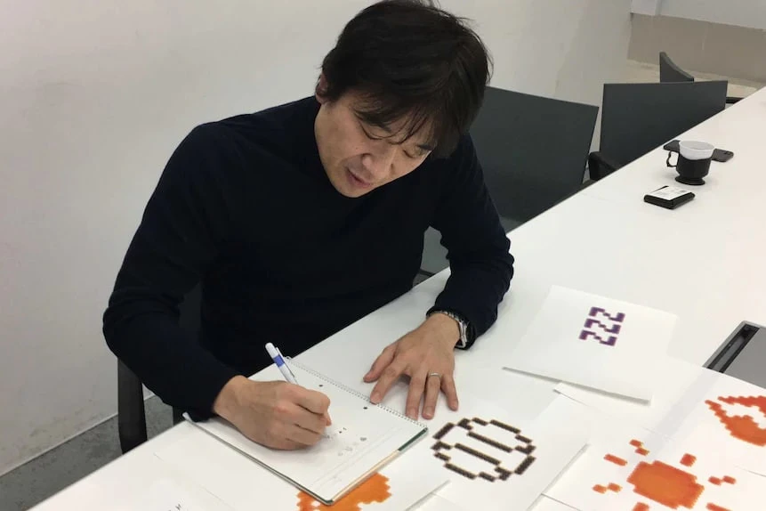 Shigetaka Kurita drawing in a notebook at a table with sheets of enlarged pixel symbols.