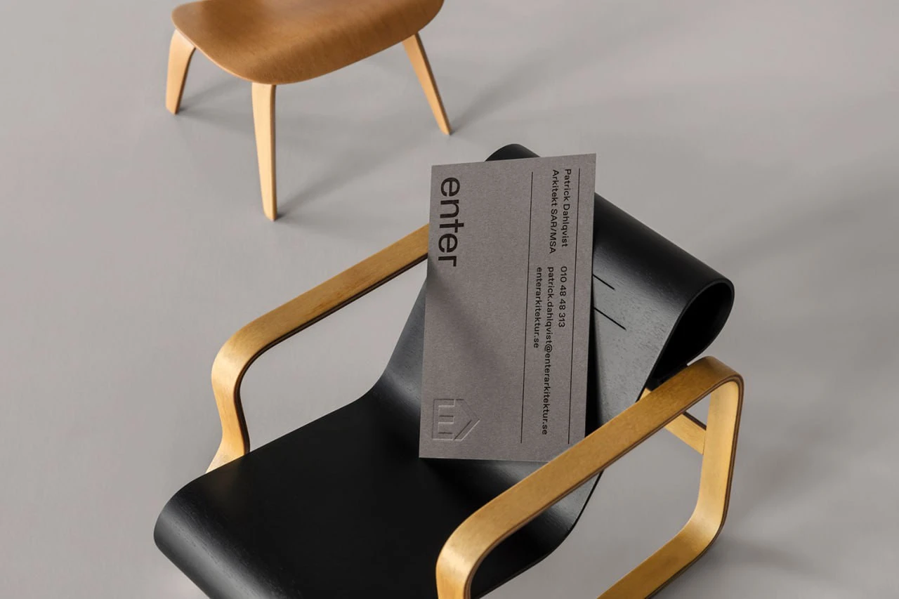 A grey Enter Arkitektur business card with black typography and an embossed mark, placed on a black chair with wooden armrests.