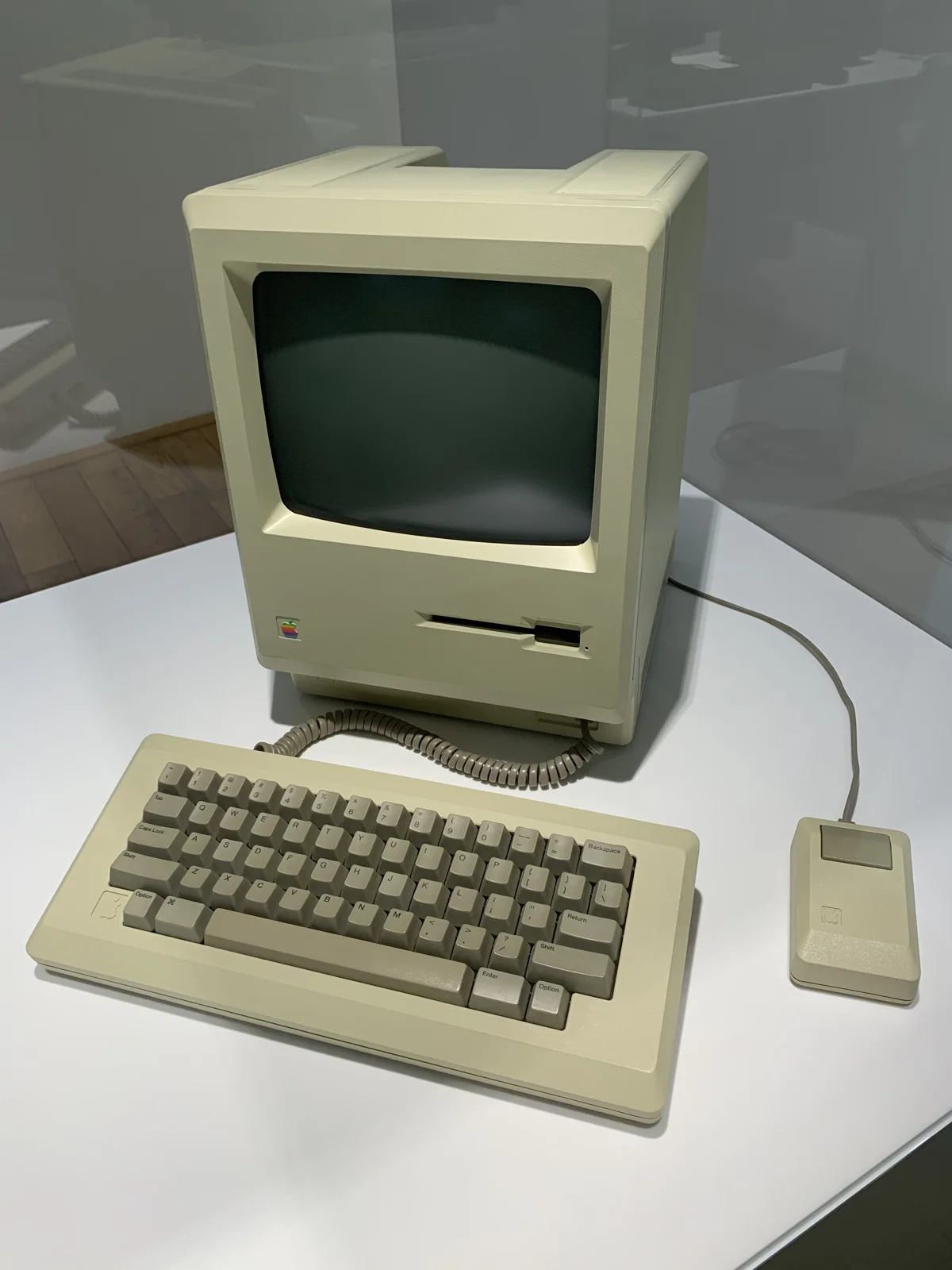 Бежевий Macintosh 128K із клавіатурою та однокнопковою мишею в музейній вітрині.