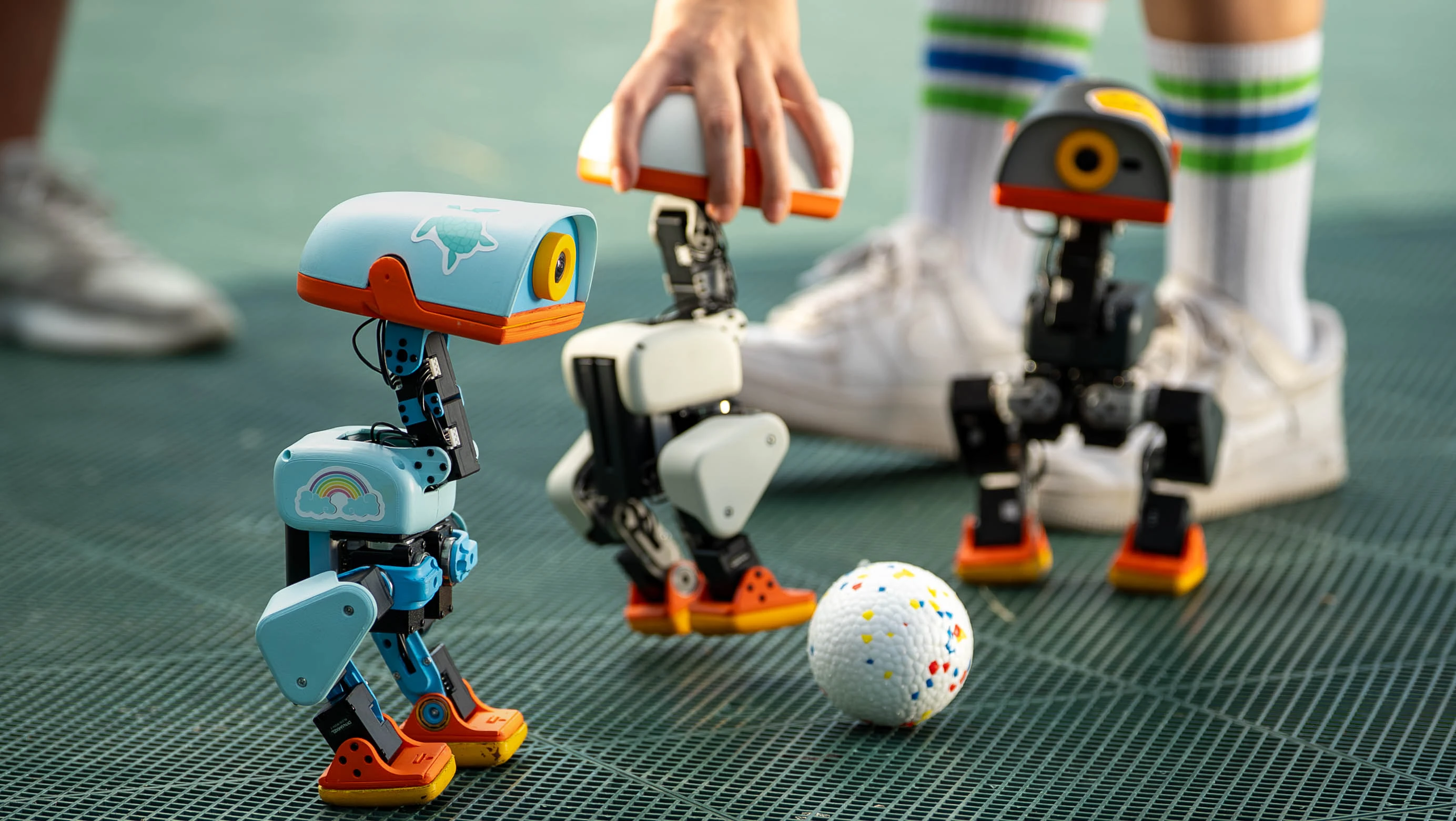 Three Microduck robots in different colours stand beside a white ball, with the blue robot closest to the camera.